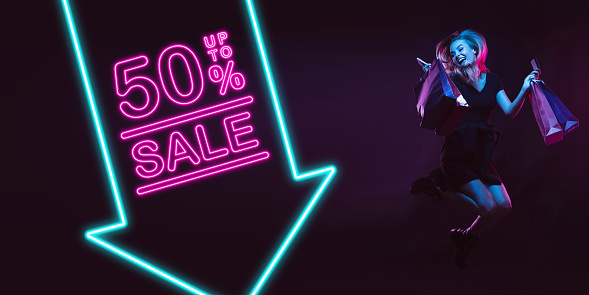 Cheerful, jumping. Portrait of young woman in neon on dark studio backgound. Human emotions, black friday, cyber monday, purchases, sales, finance concept. Copyspace. Seamless post for instagram.