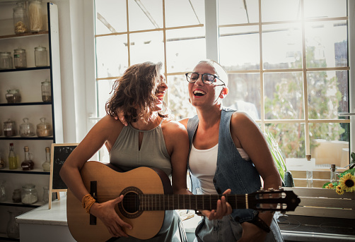 Two girl friends playing guitar in the kitchen near window at home