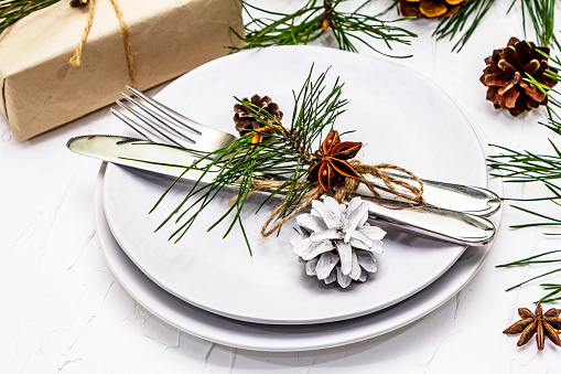 Christmas or New Year table setting. Festive cutlery, ceramic plates with fresh pine branch, cones, zero waste gifts and star anise. White putty background, close up