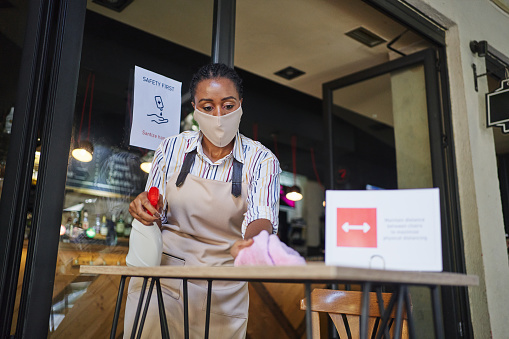 Female cafeteria worker using cleaning product and wet wipe to disinfect table at outdoor cafeteria during COVID-19 pandemic and on the table is a sign about six feet rule