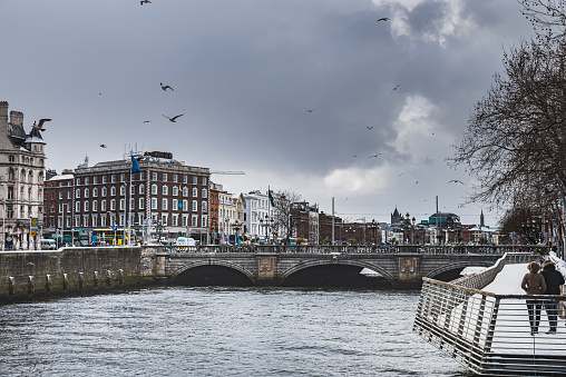 Relaxed people walk on the snowy River Liffey promenade as seagulls fly in the scene. Calm couple enjoy the grey inner city skyline with the busy O' Connell Bridge - Dublin, Ireland