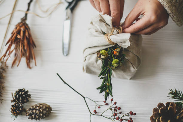 istock Female preparing zero waste christmas present. Hands decorating stylish christmas gift in linen fabric with green branch on white rustic table with natural herbs and pine cones. 1270662860