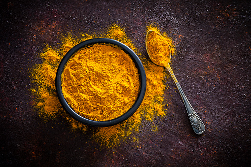 Spices: Top view of a black bowl filled with turmeric powder shot on abstract brown rustic table. A metal spoon with turmeric powder is beside the bowl and turmeric powder is scattered on the table. Predominant colors are brown and yellow. Low key DSRL studio photo taken with Canon EOS 5D Mk II and Canon EF 100mm f/2.8L Macro IS USM.