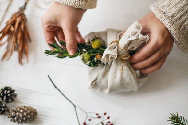 istock Hands decorating stylish christmas gift in linen fabric with green branch on white rustic table with natural herbs and pine cones. Female preparing plastic free christmas present. 1270663094