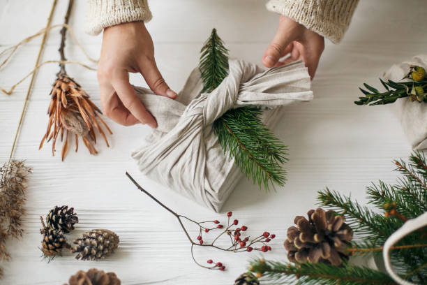 istock Florist preparing zero waste Christmas gift. Plastic free holidays. Hands decorating stylish christmas gift in linen fabric with green fir branch on white rustic table with pine cones and berries. 1270663311