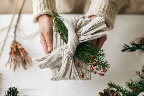 zero waste christmas holidays. hands holding stylish gift wrapped in linen fabric with green fir branch and red berries on rustic background. plastic free sustainable lifestyle - fir branch imagens e fotografias de stock