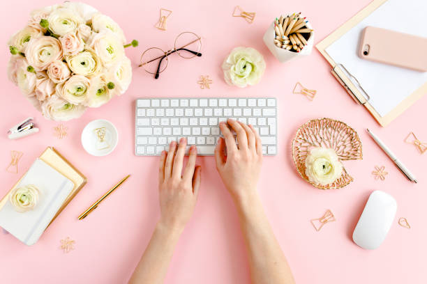 istock Stylized, pink women's home office desk. Workspace with female hands, computer, bouquet ranunculus and roses, clipboard on pink background. Flat lay 1270671559