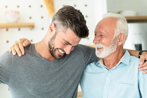 Close-up of senior man and his son embracing and laughing in the kitchen at home.