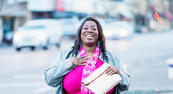 A mid adult African-American woman standing outdoors on a city street. She is smiling and looking at the camera, holding a purse.
