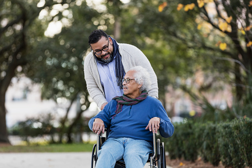 A senior Hispanic man in his 80s sitting in a wheelchair, taking a walk in the park with his adult son, a mid adult man in his 30s.