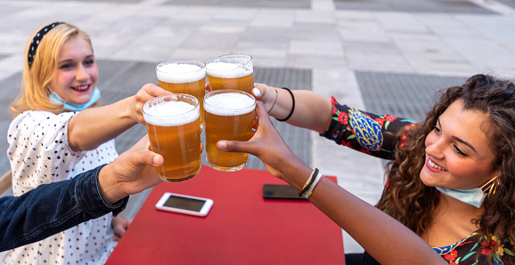 group of multi-ethnic friends toast raising glasses of beer, focus on the four raised beers and the hands of millenials toasting, blonde girl and curly brunette in the background