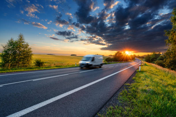 le mouvement brouillé blanc fourgon de livraison rapide conduisant sur la route d’asphalte dans le paysage rural dans les rayons du coucher du soleil avec le nuage foncé de tempête - fast motion photos photos et images de collection