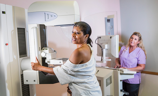 A mature African-American woman in her 40s wearing a hospital gown, getting her annual mammogram.  She is being helped by a technologist, a blond woman wearing scrubs. The focus is on the patient.