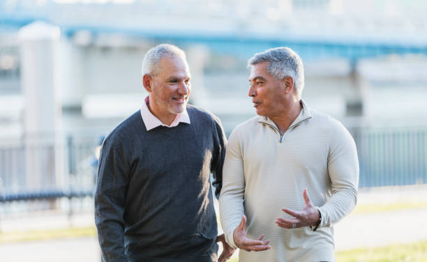Two Hispanic men conversing as they walk on a city waterfront. The are looking at each other. The one on the right is a senior man in his 60s. His friend is a mature man in his 50s.