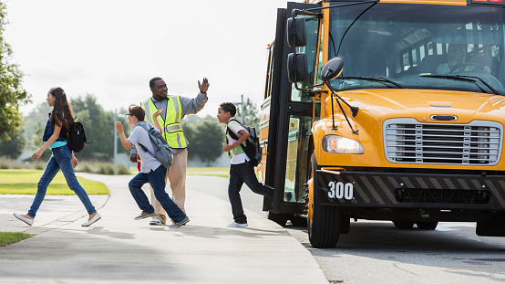 A group of three middle school students, 12 and 13 years old, exiting a yellow school bus. The crossing guard is waving to the bus driver sitting inside the bus. The boy in the middle wearing eyeglasses has down syndrome.