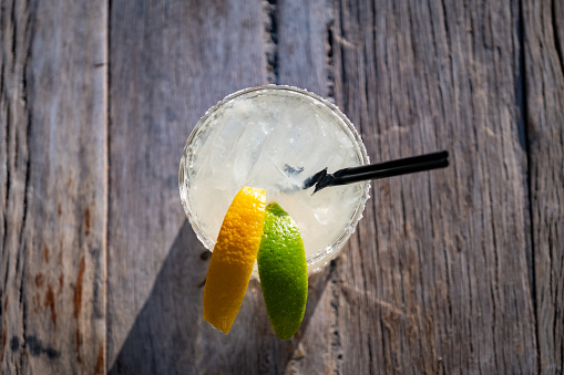 A close up looking down on a perfectly refreshing Margarita resting outside on a wood picnic table. The weathered wood of the picnic table makes a great place for copy. The drink is garnished with a lemon, and lime, and a black straw.