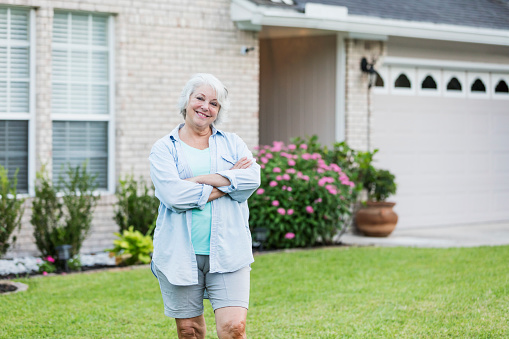 A senior woman standing in front of her house, in the  front yard. She is a happy, retired homeowner, arms crossed, smiling at the camera. She is in her 60s with white hair, wearing casual clothing.
