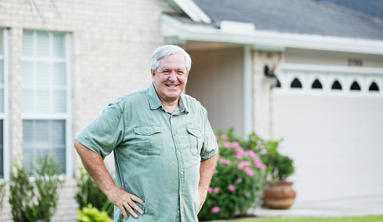 A senior man standing in front of his house, in the front yard. He is a happy, retired homeowner, hands on his hips, smiling at the camera. He is in his 60s with white hair, wearing casual clothing.
