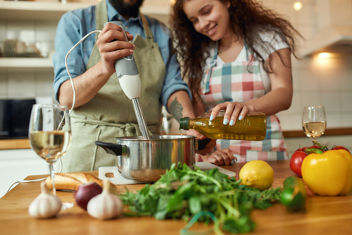 Cropped shot of man, chef cook using hand blender while preparing a meal. Young woman, girlfriend in apron pouring olive oil in the pot, helping him in the kitchen. Cooking at home. Horizontal shot
