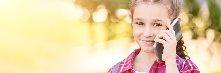 Cute little girl talking. Hold phone near face. Young woman smile. Looking at camera. Outdoor school. Green background. Smiling speach. Happy expression. Student lifestyle. Horizontal banner