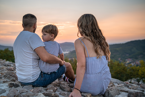 Young parents and their little son are sitting on the stone wall and enjoying the beauty of nature.