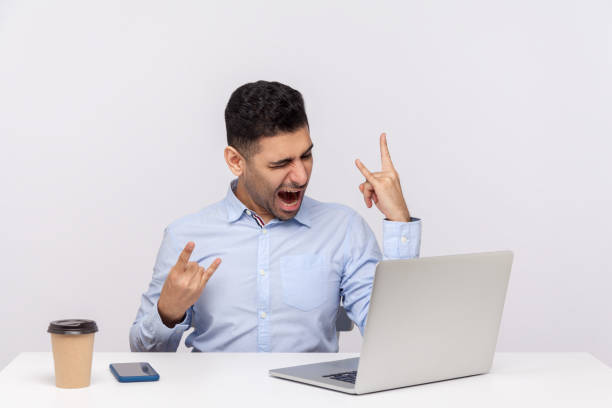 Carefree joyful businessman sitting office workplace, showing rock and roll hand gesture, devil horn sign