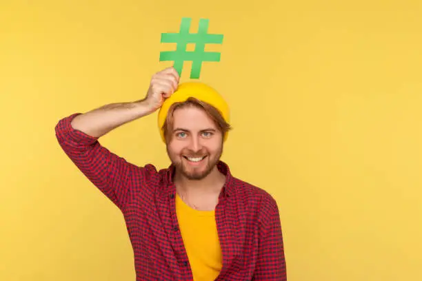 Photo of Hashtag, internet trends. Happy funny hipster guy in checkered shirt smiling and holding hash sign over head