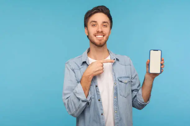 Photo of Portrait of cheerful happy man in worker denim shirt smiling and showing mobile device, cell phone with mock up