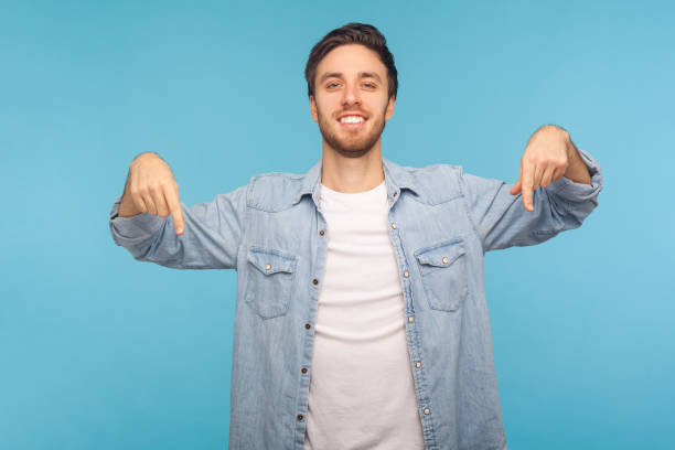 Look at ads below! Portrait of handsome happy man in stylish denim shirt smiling and pointing down Look at ads below! Portrait of handsome happy man in stylish denim shirt smiling and pointing down, showing place for idea presentation, commercial text. indoor studio shot isolated on blue background man hand down stock pictures, royalty-free photos & images