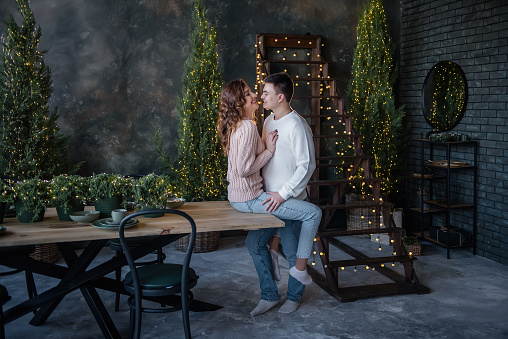 A girl in a pink knitted sweater sits on a wooden table, she is hugged by a young man in white. A loving couple hugs, kisses on New Years Eve. The background of garlands of lights, Christmas trees