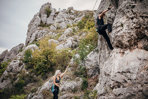 Man and woman, male rock climber is climbing on rocks in nature, woman is assisting him by holding the rope.