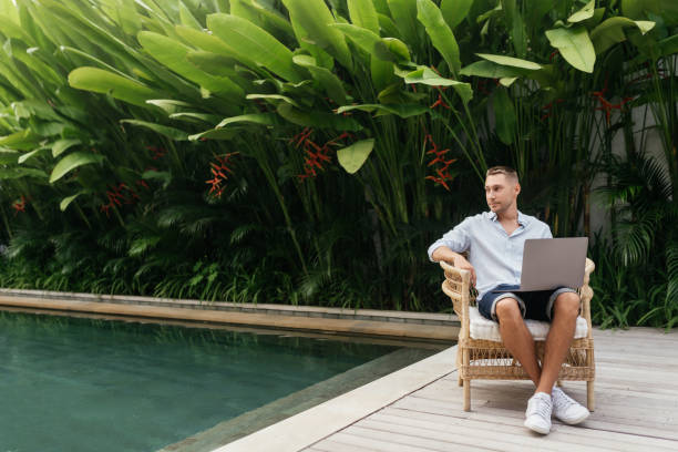 istock Young man using a laptop computer in a garden with a swimming pool. 1270773010