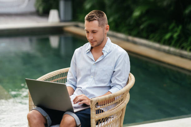 istock Young man using a laptop computer in a garden with a swimming pool. 1270773014