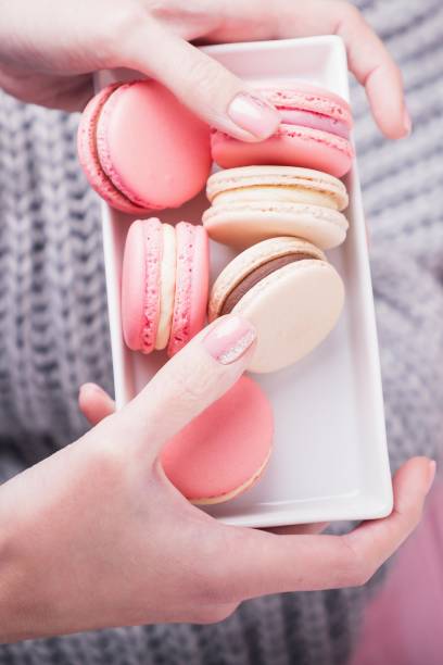 istock Female hands holding rectangular white plate with delicious pink and beige macaroons on pink and gray background. Top view, copy space 1270779938