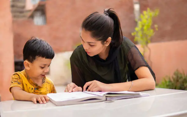 Outdoor image of Indian young girl tutor teaching little boy. They are sitting together at study table on rooftop in day time.