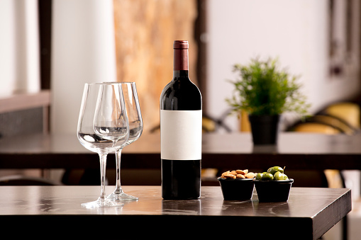 Wine bottle with white label for text with wine glasses and olives and almonds, on top of the wooden table, inside the restaurant and free space for decoration.