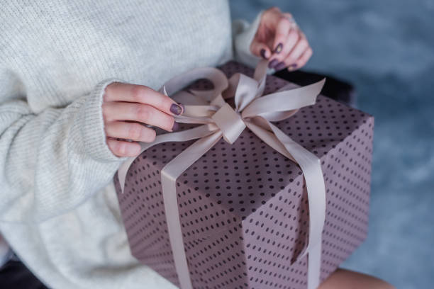 Female hands hold powdery gift ribbons. The box is packed in purple paper, lying on the lap of the girl in a warm knitted sweater with burgundy manicure. Christmas surprise for birthday. Post card las manos femeninas sostienen cintas de regalo polvorientos. la caja está embalada en papel púrpura, acostada en el regazo de la niña en un suéter de punto cálido con manicura burdeos. sorpresa de navidad para el cumpleaños. tarjeta postal
