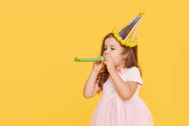 Photo of Party time. A joyful little girl in a festive cap and elegant dress celebrates her birthday. Blowing a whistle on a yellow background