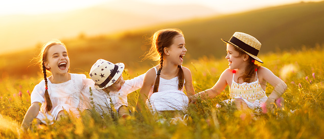Cheerful little kids in casual white clothes sit in the grass, having fun and laugh merrily  in together in field at sunset while enjoying summer holidays in countryside