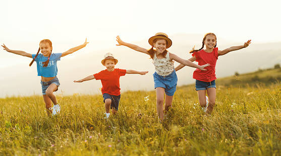 Cheerful little kids in casual clothes having fun and running back in together in green field at sunset while enjoying summer holidays in countryside