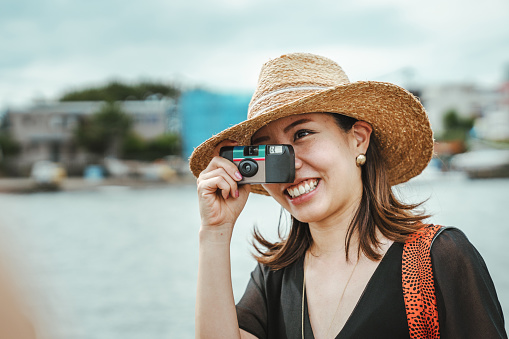 A side shot of a smiling Asian woman wearing a straw hat and using a one-time-use camera to take city photo outdoors next to the sea