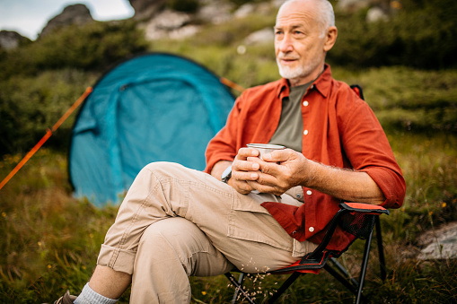 Active Senior Man Camping in Mountains. Sitting in a front of his Tent on Camping Chair, Enjoys a Cup of Coffee