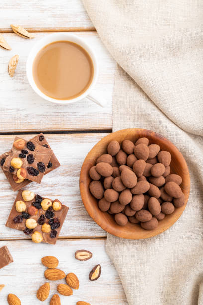 amande dans des dragées de chocolat dans l’assiette en bois et une tasse de café sur le fond en bois blanc et le textile de lin. vue supérieure, gros plan, plat. - chocolate almond foods and drinks white chocolate photos et images de collection