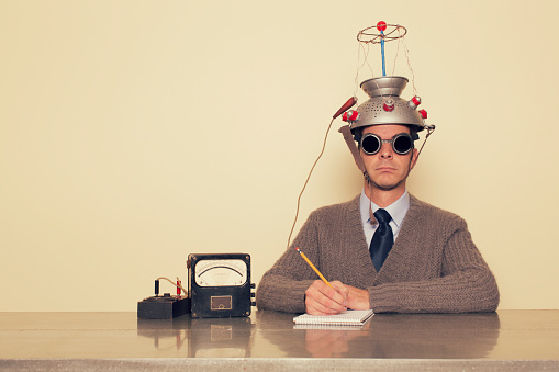 A man is testing the limits of the mind by placing a mind reading invention on his head trying to understand the brain. He is dressed in retro sweater and tie with safety goggles waiting to measure brain waves and understand the unknown.