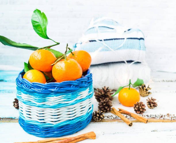 christmas basket with tangerines, cinnamon sticks and pine cones lie on a wooden white background. knitted jumpers stack on top of each other and the garland burns beautifully. hygge concept - m9 imagens e fotografias de stock