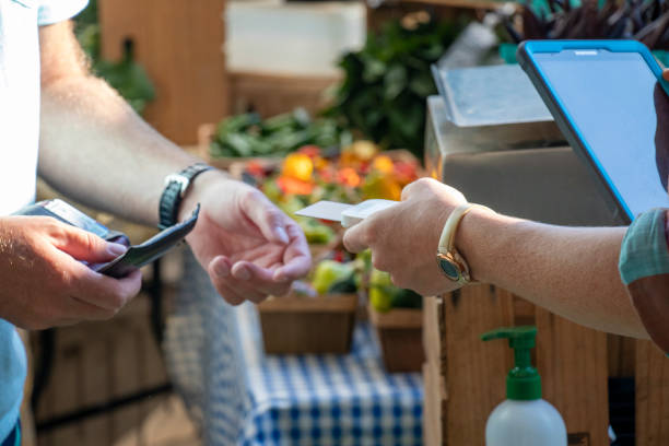 A vendor holding an electronic tablet accepts a credit card payment using a touchless card reader at an outdoor market. stock photo