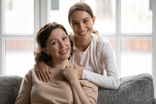 Portrait of smiling millennial woman cuddling happy beautiful middle aged mother sitting on sofa. Happy multigenerational female loving family looking at camera, posing for photo together at home.