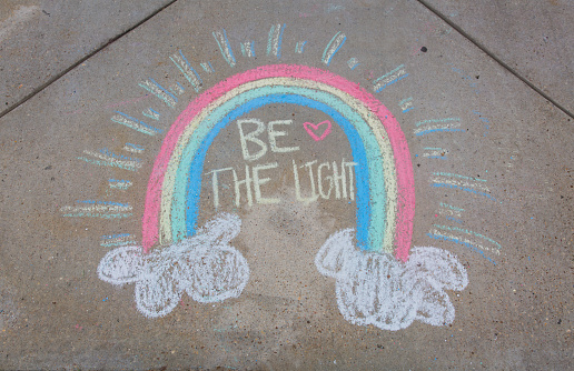 Be the light written with sidewalk chalk with a rainbow