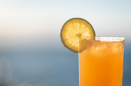 A close up of a perfectly refreshing Mai Tai resting outside on a chair by the side of a lake. The gradient blues of the lake and the sky above the horizon are soft and out of focus in the background. The Sun is setting on a cloudless day and the sunlight reflects off the water. The drink is garnished with a lime wheel.