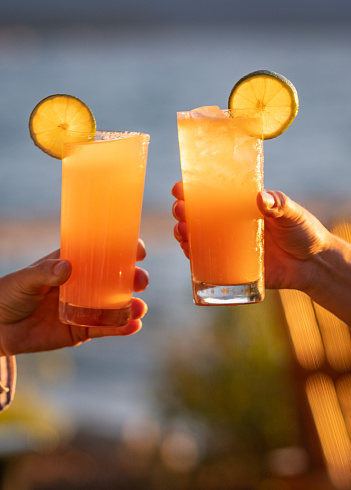 A celebratory toast lakeside at sunset with two orange cocktails on the rocks (could be Mai Tai's, Tequila Sunrises, Screwdrivers, or any number of drinks) garnished with lime wheels.